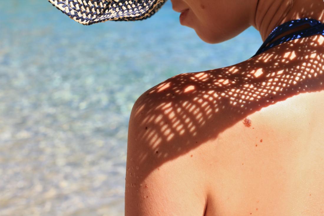 Mulher tomando banho de sol pelo mar, usando chapéu para proteger a pele.