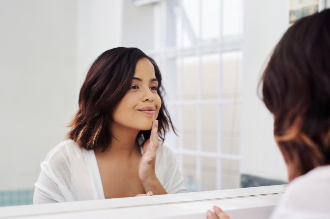 Жена гледа в банята огледало и се усмихва, докато овлажнява лицето си.