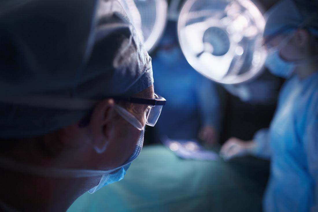 Хирурзи, работещи в операционната зала, силует на хирург, носещ очила и маска на преден план.