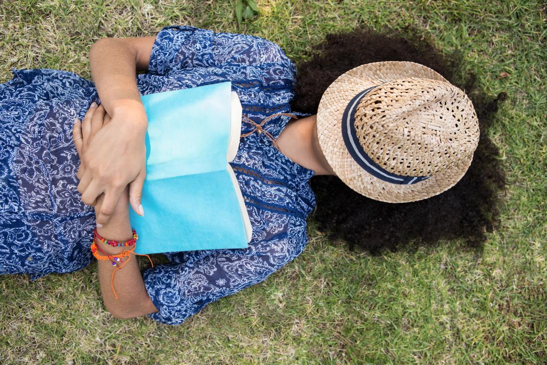 Жена лежаща на трева с книга и шапка, покриваща главата