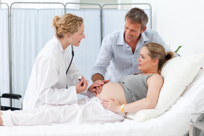Бременна жена, лежаща в леглото, говори с лекар и партньора си.