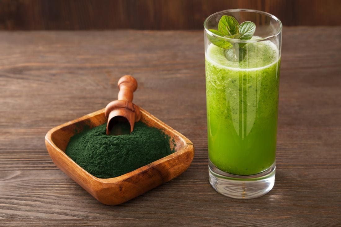 Pó verde do chlorella em uma bacia de madeira, ao lado de um vidro com uma bebida verde do smoothie.