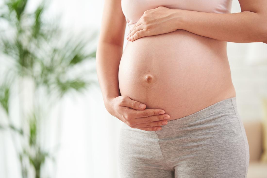 Schwangere Frau, die ihren ausgesetzten Bauch mit Anlage im Hintergrund golding ist.