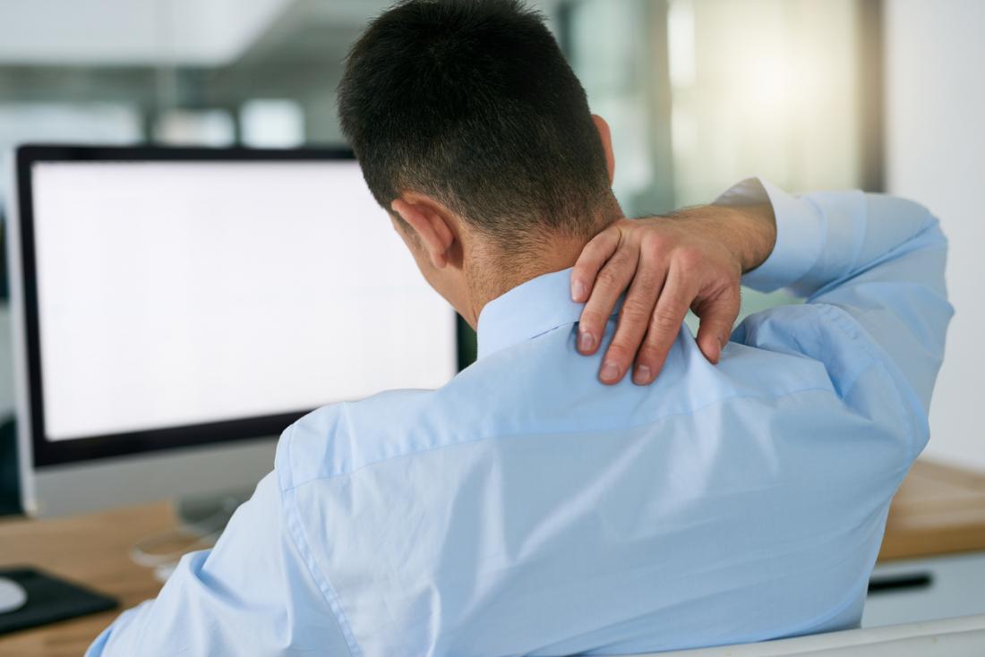 Човек в работно облекло, седнал на бюрото пред компютъра, държейки гърба на врата си в болка.