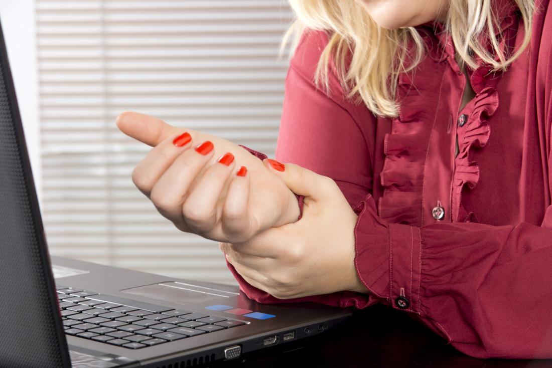 Жена, работеща върху лаптоп, която държи китката си, поради повтарящи се наранявания.