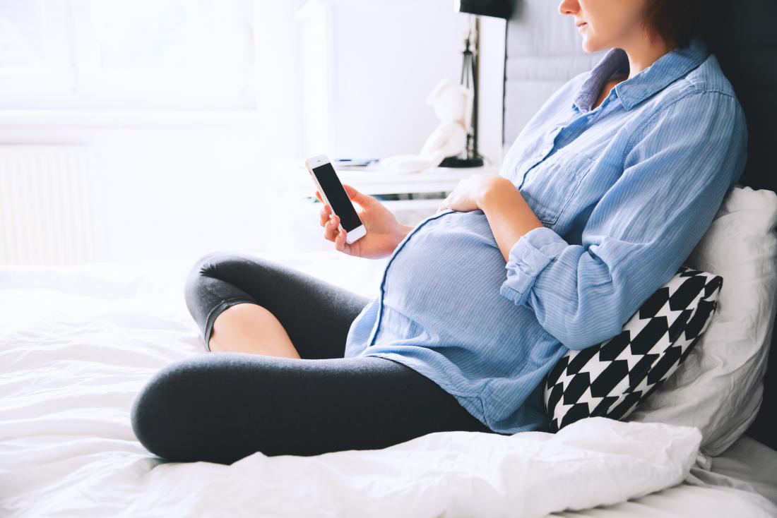 donna incinta che si siede sul letto usando smartphone