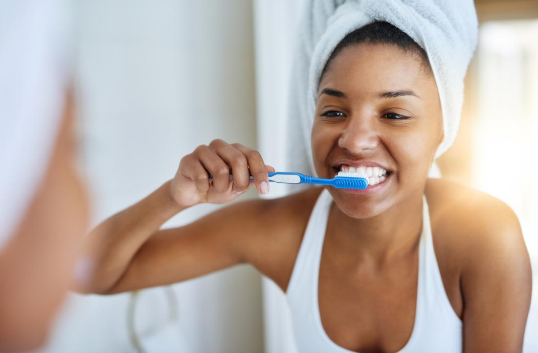 Mulher escovando os dentes na frente do espelho do banheiro, o cabelo enrolado em uma toalha.