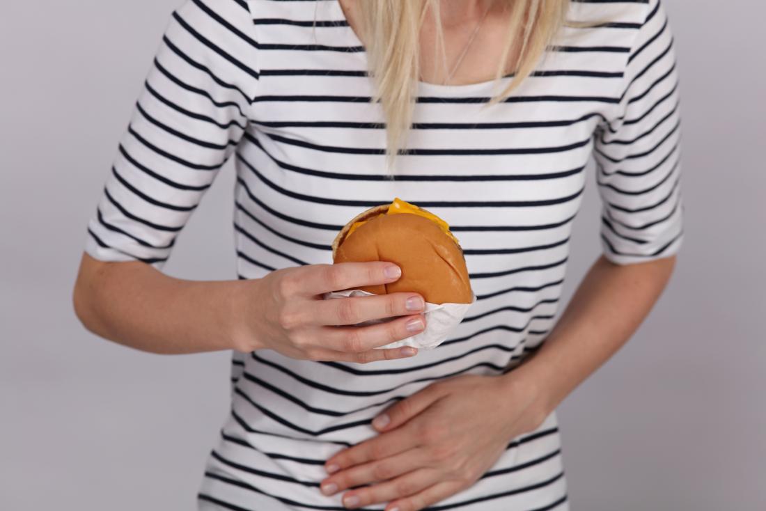 Жена със стомашна болка след ядене, държаща бургер и ръка пред корема си.