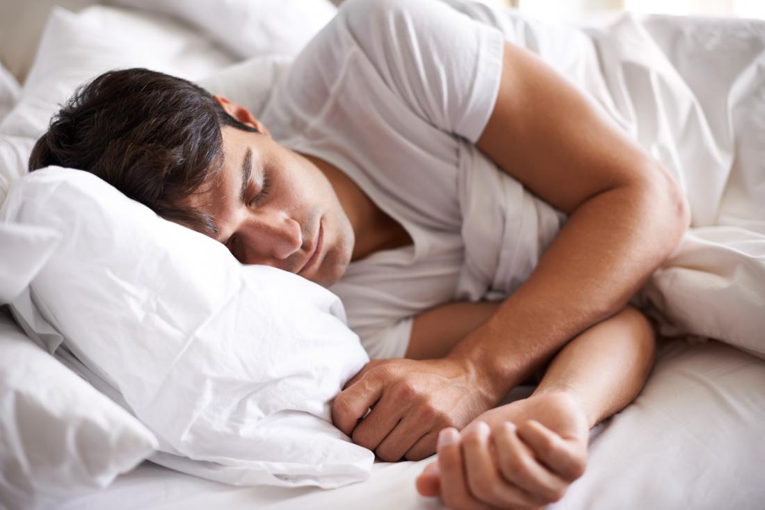 Човек в бяла тениска, заспала в леглото с бели чаршафи.