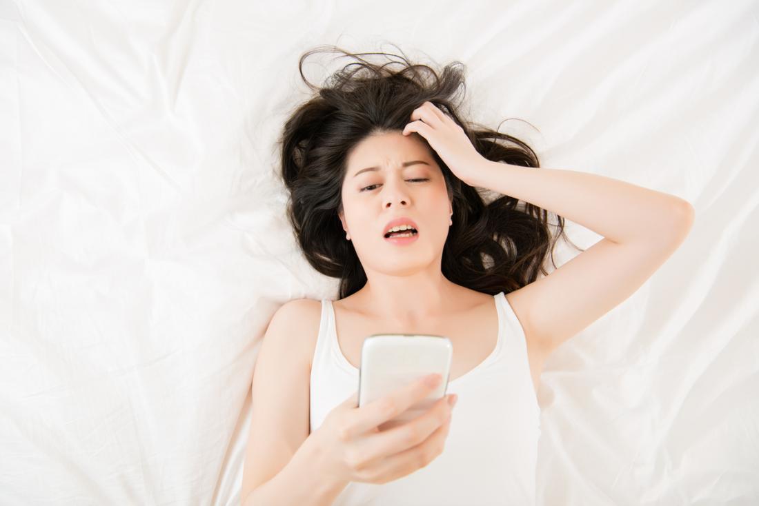 Frau mit den Kopfschmerzen, die auf Bett legen und Telefon betrachten